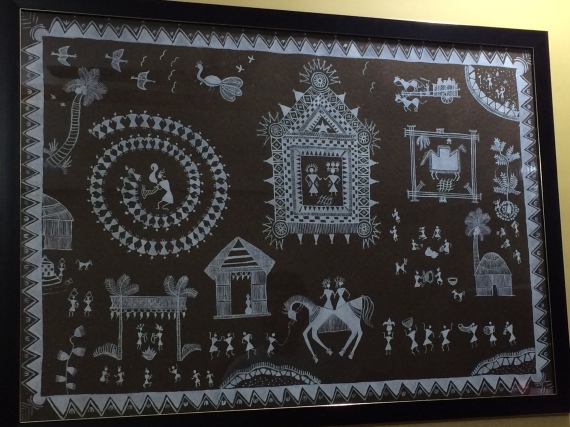 Warli Painting showing temple in center, people dancing, marriage ceremony, men working in fields, women gathered around weaving baskets, children playing and various other daily activities.